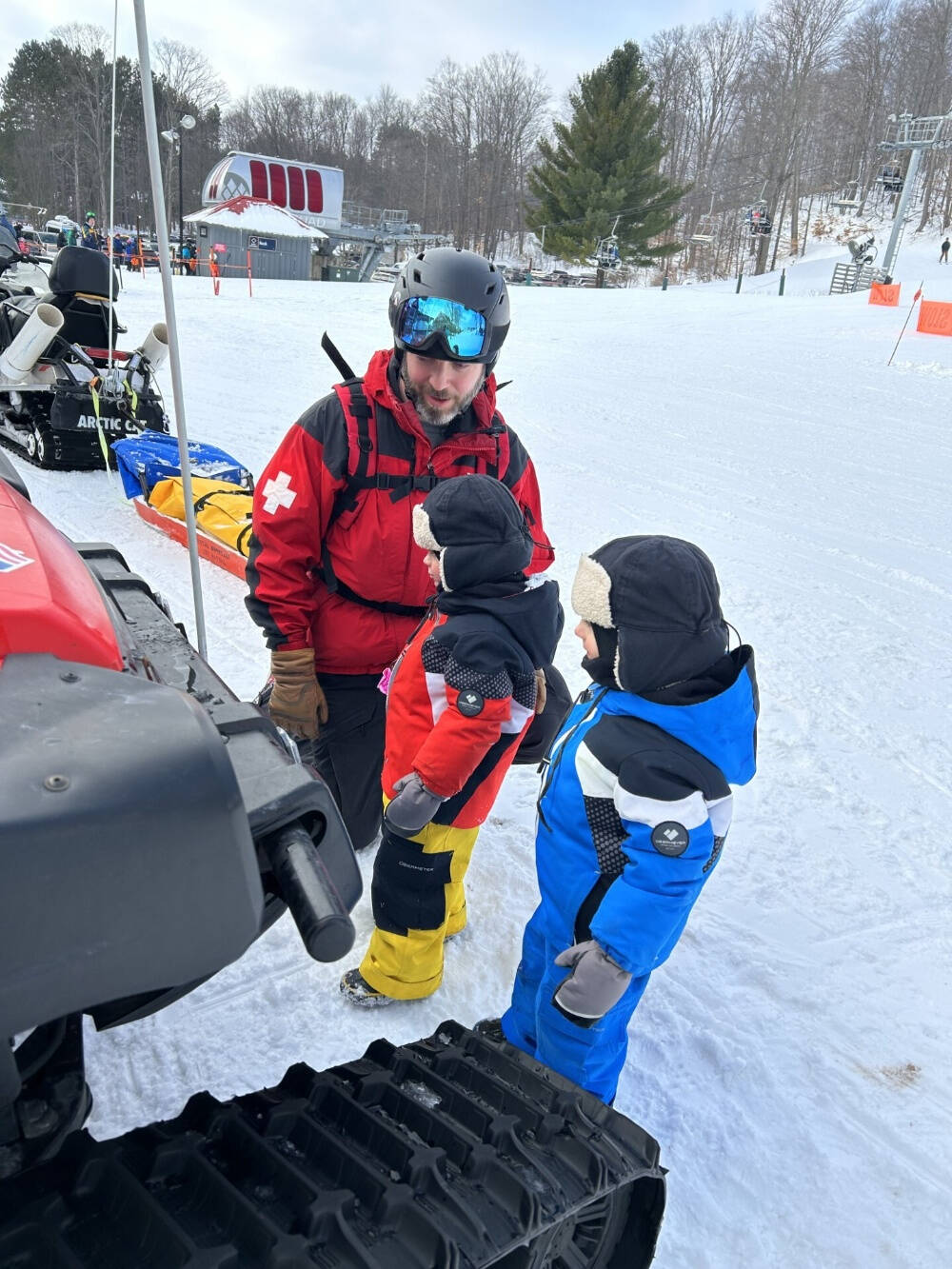 Ski patrol member Patrick Smith speaking to two children on a snowy ski slope near a snowmobile.
